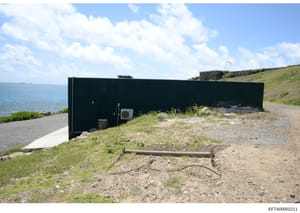 This photograph shows a modern black rectangular structure built into a coastal hillside overlooking the ocean. The building appears to be a utility or service facility with minimal visible features, situated on a grassy slope with a flagpole visible