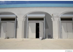This photograph shows the exterior of a white concrete building structure located on a beach, featuring multiple arched entrances with white doors. The building has a simple, modern design with a turquoise roof visible at the top. The image appears t