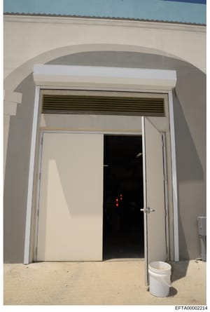 This photograph shows an arched interior doorway with white trim and a closed white door. The door is partially open, revealing a darkened interior space with red lights visible inside. A white bucket is positioned on the sandy floor in front of the 