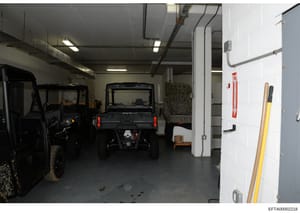 This is a photograph of an indoor garage or storage facility containing multiple utility vehicles including what appears to be ATVs or small off-road vehicles, stacked hay/straw, and various tools and equipment. The space has industrial lighting, con