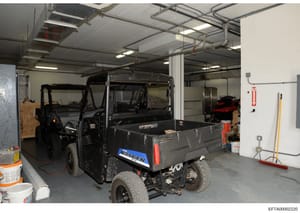 Photograph of an industrial or commercial garage space showing multiple utility vehicles including a Ranger-branded utility cart and other equipment. The space appears to be a storage or maintenance facility with fluorescent lighting, white walls, an