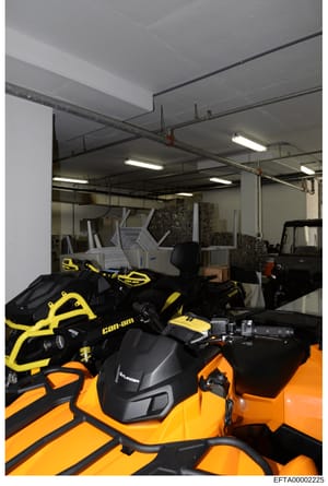 Photograph of an indoor storage or warehouse facility containing multiple all-terrain vehicles (ATVs), including Can-Am branded models in yellow and black, and orange Polaris vehicles. The space also contains stacked materials, white equipment, and v