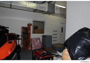 This photograph shows an interior storage or basement space containing various items including wrapped furniture, storage boxes, tools, and equipment. The room has white cinder block walls, exposed ceiling infrastructure, and fluorescent lighting. Th