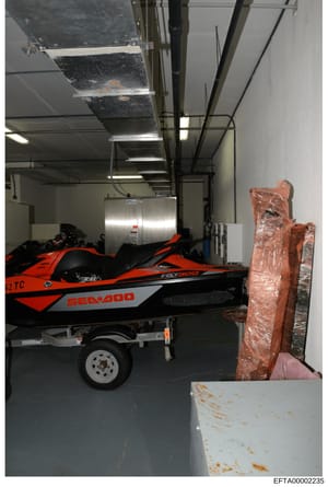 This is an evidence photograph showing the interior of a storage or maintenance facility containing a red and black Sea-Doo jet ski on a trailer, along with what appears to be wrapped furniture or items. The image documents the condition and location