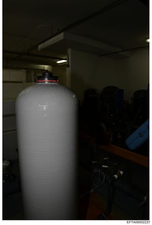 Photograph of a large white cylindrical storage tank or water filtration system in an indoor facility. The equipment appears to be industrial or commercial in nature, featuring a red valve assembly at the top and black piping connections. The image w