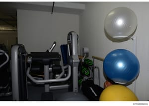 This is a photograph of a residential gym or fitness room showing various exercise equipment and accessories. The image captures exercise machines, stability balls, dumbbells, and other workout gear stored in what appears to be a basement or dedicate