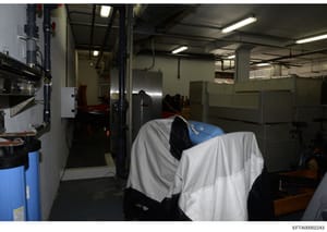 This photograph shows an industrial or basement-level space with utility equipment, mechanical systems, and storage areas. The image captures what appears to be a service or mechanical room with fluorescent lighting, exposed piping, metal storage con
