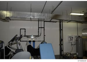 This photograph shows an interior room with exposed mechanical systems including ductwork, piping, and HVAC equipment mounted on walls and ceiling. A blue piece of exercise or medical equipment is visible in the foreground. The image appears to be do