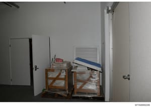 This photograph shows a storage or utility room containing two wooden-framed crates with plastic-wrapped contents and various items stacked on top, including what appears to be a white vent cover and other equipment. The room has plain white walls, a