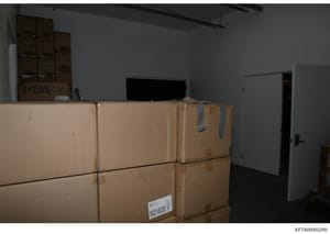 This photograph shows an interior storage or warehouse space containing multiple sealed cardboard boxes stacked against a wall, with shelving units visible in the background and a television monitor on top of boxes. The image appears to be evidence d