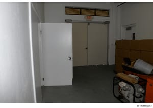 This is a photograph of an interior room or basement space, showing white doors, fluorescent lighting, an EXIT sign, and various equipment and storage items including boxes and tools. The image appears to be evidence documentation from a property sea