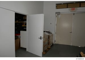 This is an evidence photograph showing an interior room or corridor with an EXIT sign visible on the wall. The space contains white doors, storage equipment including what appears to be a wrapped or covered item on the floor, and various other materi