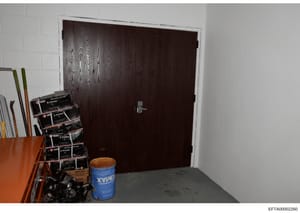 This is an evidence photograph showing an interior room or storage space containing various items including stacked clamps, tools, and a blue container labeled 'XYPEX'. The room features white painted block walls, a dark wood double door with a handl