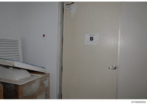 This is an evidence photograph showing the interior of a room, likely from a property search or seizure. The image displays a door marked with an evidence label 'B', a wall-mounted ventilation unit, an electrical outlet, and what appears to be wooden
