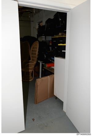 This is an evidence photograph showing the interior of a storage closet or small room containing various stored items including wicker chairs, luggage, black equipment or storage boxes, and miscellaneous household items. The photo documents the conte
