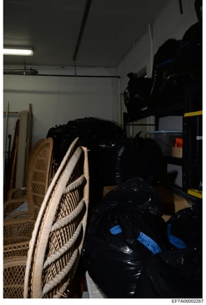 This is an evidence photograph showing an interior storage space containing multiple sealed black plastic bags or containers labeled with blue tape markings, woven rattan chairs, rolled materials, and various storage items. The sealed containers and 