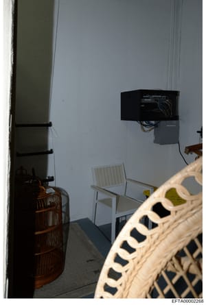 This is an evidence photograph showing the interior of a room containing electrical wiring, a black control box mounted on the wall, white chairs, and various items including what appears to be a decorative birdcage. The image documents physical cond