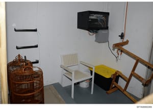 This is an evidence photograph showing an interior room containing various items including electronic equipment mounted on the wall, wooden furniture, chairs, and storage containers. The photo appears to be from a property search or inventory documen