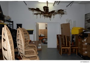 This photograph documents a storage or evidence room containing furniture and items, likely seized during the investigation. The image shows various pieces of furniture including wicker chairs, wooden dining chairs, cardboard boxes, and a distinctive