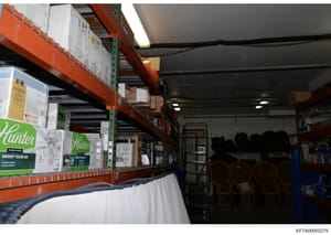 This is a photograph of a storage warehouse or inventory facility showing industrial shelving with orange and green support beams containing various boxed items and supplies. The image appears to be documentary evidence, possibly related to property 