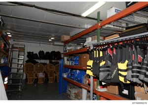This is a photograph of an industrial warehouse or storage facility containing various equipment and supplies. The image shows organized storage with orange industrial shelving units holding life jackets, wetsuits, helmets, paddles, and other water s