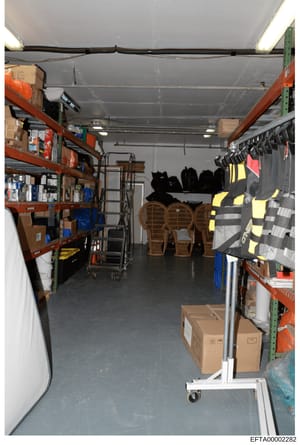 This photograph shows the interior of a storage garage or warehouse space containing organized shelving units, miscellaneous supplies, and equipment. The space includes red and green industrial shelving units stocked with boxes and supplies on the le