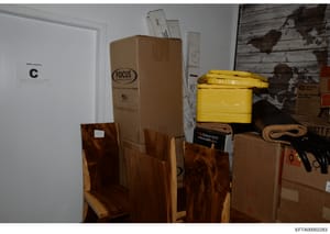 This is a photograph of a storage or utility room showing various items including cardboard boxes, wooden furniture pieces, a yellow plastic container, tools, and other household materials. The image appears to be documentation of property contents, 