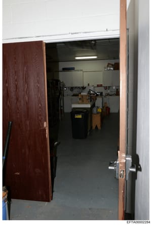 This photograph shows the interior of a storage or utility room, featuring dark flooring, white walls with shelving units, and various equipment and boxes stored inside. The room appears to be accessed through a dark wood-framed doorway. The image wa