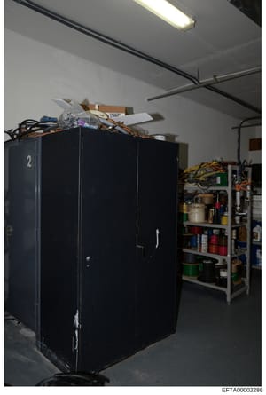 This is an evidence photograph showing a storage or utility room containing a large black numbered cabinet (marked with '2'), metal shelving units with various supplies and materials, electrical wiring, and miscellaneous boxes and items on top of the