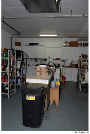 This is a photograph of an industrial or residential garage/storage space containing various tools, equipment, and supplies organized on shelves and work surfaces. The image shows a work area with metal shelving units, cardboard boxes, a black waste 