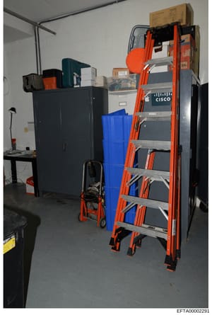 This photograph documents the interior of a storage or equipment room containing various items including a tall orange ladder, blue plastic containers, black filing cabinets, boxes, and networking equipment with visible Cisco branding. The image appe