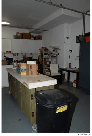 This is an evidence photograph showing an interior storage area or basement space containing various cardboard boxes, wooden furniture, shelving units, and miscellaneous items. The room appears to be used for storage purposes and may be from one of t