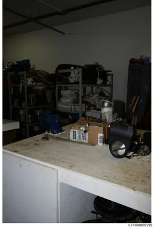 This is an evidence photograph showing an interior room containing various electronic equipment, networking hardware, boxes, tools, and storage items. The room appears to be a technical or server space with metal shelving units holding computer equip