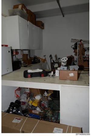 This is an evidence photograph from the Epstein investigation showing an interior storage or utility space filled with various tools, equipment, cardboard boxes, and household items. The space appears to be a garage, basement, or storage room with wh