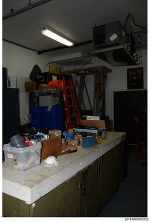 This is an evidence photograph showing the interior of a storage or maintenance room, likely from an Epstein property. The image captures various items stored on shelves and work surfaces, including tools, containers, cleaning supplies, and household