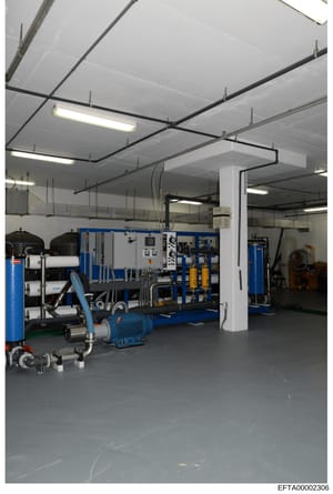 This photograph shows an industrial or commercial facility interior with specialized water treatment or processing equipment. The space contains blue-colored machinery, control panels, filtration systems, and various technical equipment arranged in a