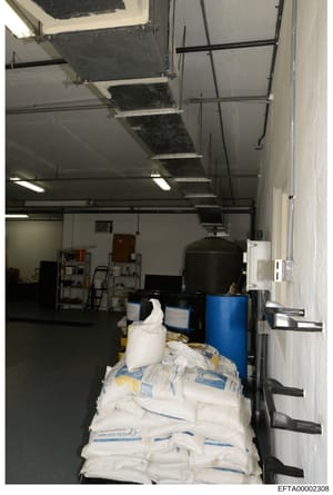 This photograph shows an interior storage or utility space, likely part of a larger facility, containing various supplies including large white bags (possibly containing bulk materials), blue drums/containers, shelving units, and HVAC equipment. The 
