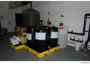 This photograph shows an indoor storage and laboratory space containing various chemical containers and water treatment equipment. Multiple large black storage drums labeled 'Sodium Hypochlorite' are visible on a yellow spill containment platform, al