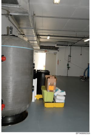 This photograph shows an industrial or commercial storage room containing large storage tanks, HVAC equipment, and various supplies in yellow containment bins. The image appears to be evidence documentation from a property search or inventory related