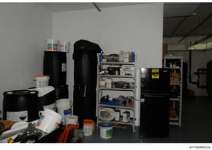 This photograph shows an interior storage or maintenance space containing various chemical containers, cleaning supplies, tools, and equipment organized on shelves and the floor. The image appears to be evidence documentation from a property search o