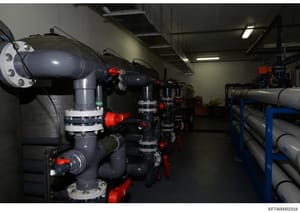 This is a photograph of an industrial facility showing multiple large dark gray/navy pipes with red valve controls and white flanged connections arranged in a warehouse or mechanical room setting. The equipment appears to be part of a water treatment