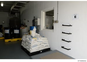 This is an evidence photograph showing the interior of a room or storage space labeled 'E', containing industrial equipment, chemical storage containers, and supplies. The image appears to be from a forensic documentation or property inventory relate