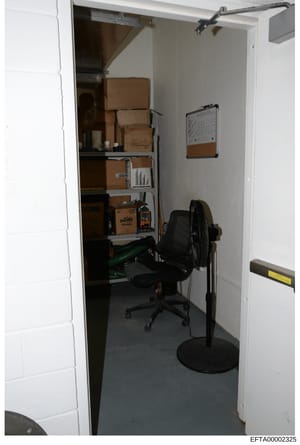 This is a photograph of a small storage closet or utility room containing cardboard boxes stacked on shelves, office furniture including a black desk chair and standing fan, and various stored items. The image appears to be evidence documentation fro