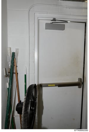 This is an evidence photograph showing an interior utility or storage space with a white brick wall, a metal emergency exit door with push bar hardware, and various stored items including cleaning tools, a fan, and maintenance equipment leaning again
