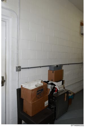 This is an evidence photograph from the Epstein investigation showing an interior room with white brick walls, a metal rod/rail system, and several brown cardboard storage boxes with labels. The purpose and specific location of this room are not imme