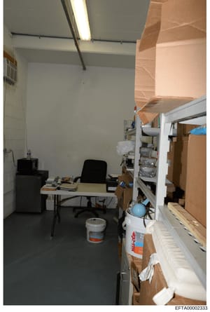 This is an evidence photograph showing an interior room with minimal furnishings and extensive cardboard boxes and storage materials. The room appears to be an office or storage space containing a desk, black office chair, filing cabinets, shelving u