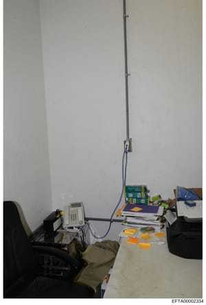 This is an evidence photograph showing an interior office or workspace containing various electronic equipment, documents, and materials. The image captures a desk area with a telephone/communications device, stacks of papers and files, storage boxes