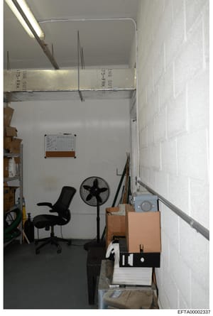 This is an evidence photograph showing an interior room with storage boxes, office furniture, and various items. The image appears to be taken during a property search or investigation, with labeled boxes visible at the top of the frame bearing marki