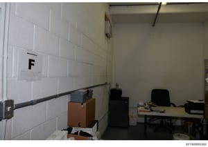 This is a photograph of a plain, institutional room with white painted brick walls. A label marked 'F' is visible on the left wall, suggesting this may be an evidence storage, archival, or office space. The room contains basic furniture including a d