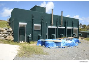 This photograph shows a dark green, industrial-style building with a flat roof, multiple white-framed windows, and metal piping. Blue tarps cover equipment or materials in front of the structure. The building appears to be located in a coastal or isl